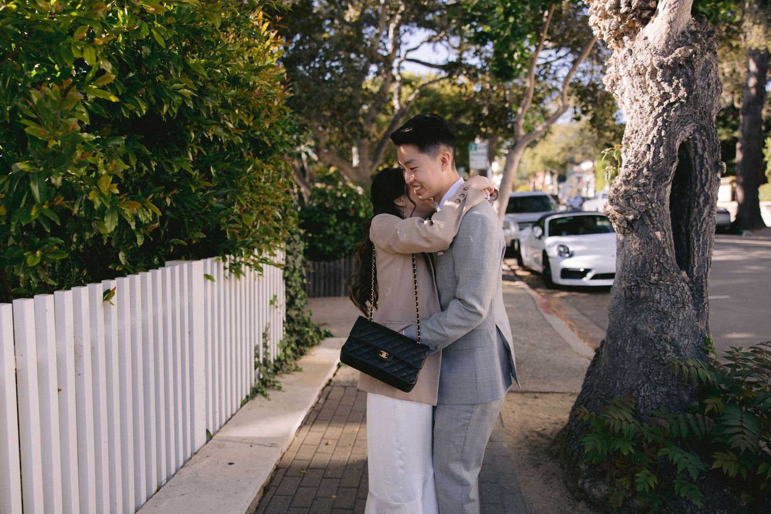 A warm embrace between two people in light-colored clothing on a sidewalk with a white picket fence and parked cars.