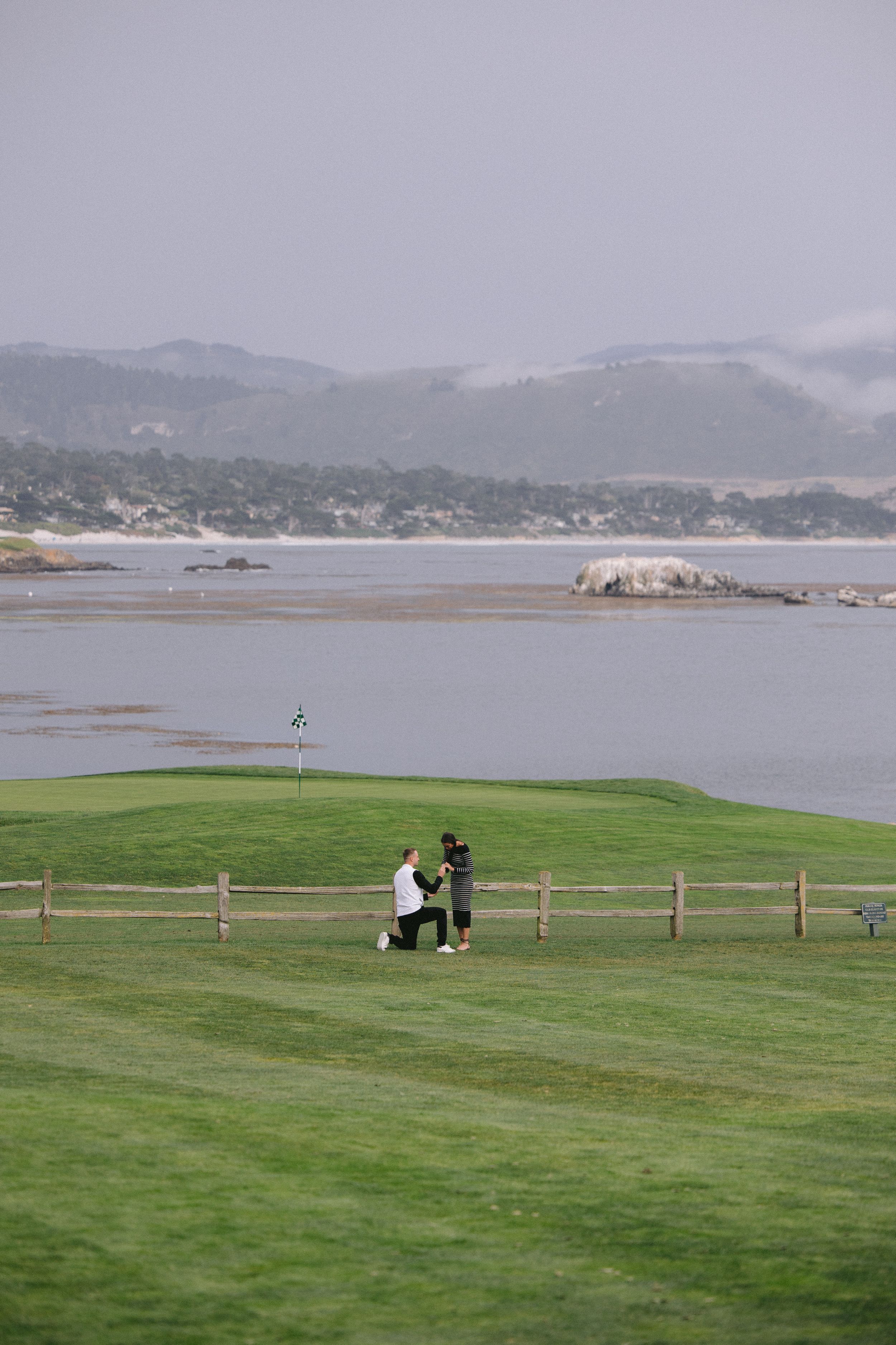 Two people standing on a coastal golf course with dramatic views of a misty bay and mountains in the background.