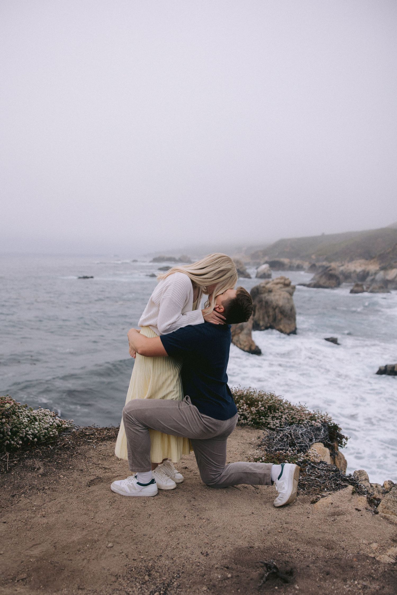 A couple embraces in a romantic moment on a foggy cliff overlooking the rocky Pacific coast.
