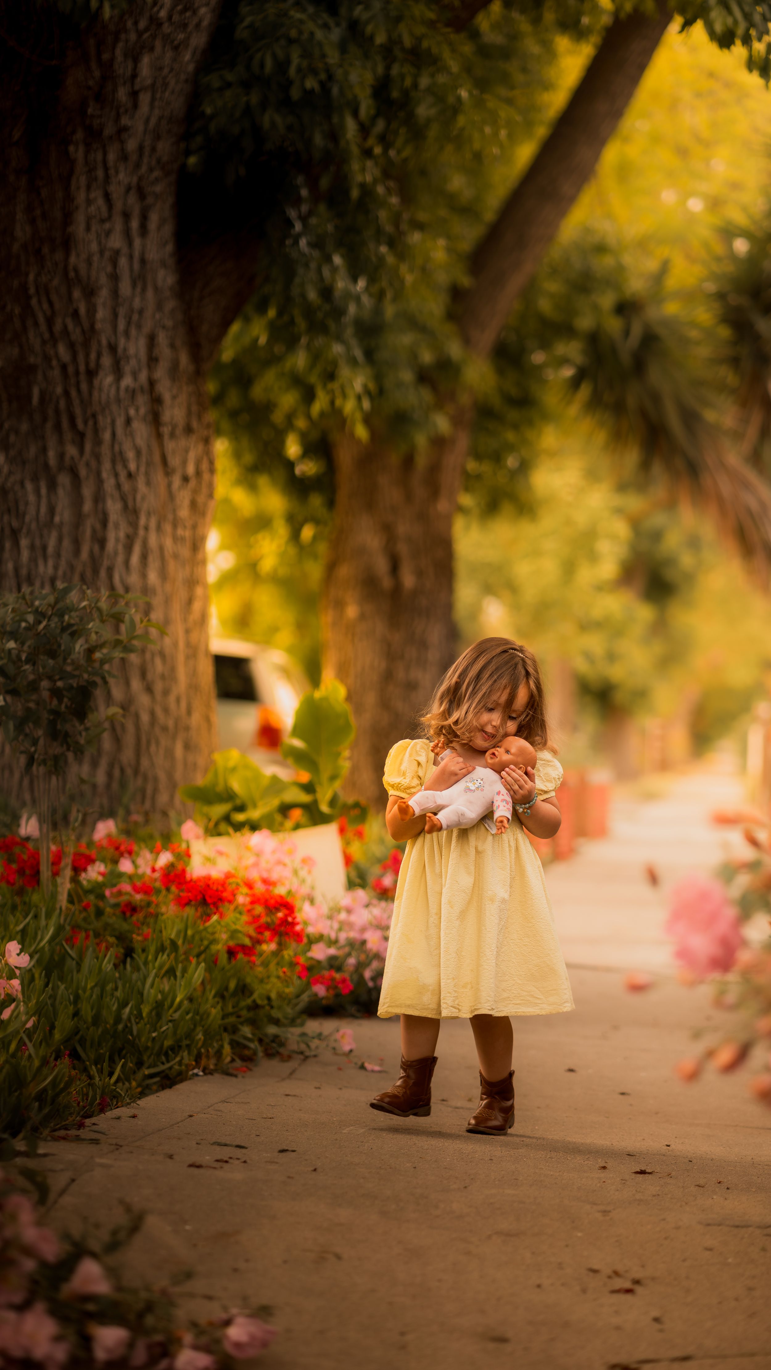 Small figure in a yellow dress and boots stands on a garden path with colorful flowers and trees in soft sunlight.