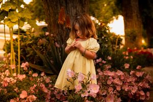 A child in a yellow dress stands among pink flowers in a dreamy garden setting during golden hour.