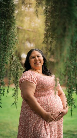 A pregnant woman in a pink floral dress stands beneath drooping willow branches on a sunny day.