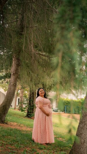 A pregnant person in a flowing pink dress stands under drooping pine trees in a peaceful garden setting.