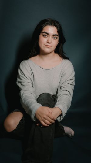 A person wearing a light gray sweater sits against a dark background in a moody portrait setting.