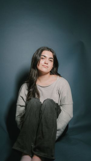 Figure sits against dark wall wearing grey sweater and dark pants in moody portrait setup.