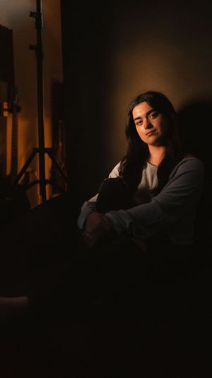 A dimly lit portrait in warm tones shows a figure in casual clothing seated against a dark background with dramatic shadows.