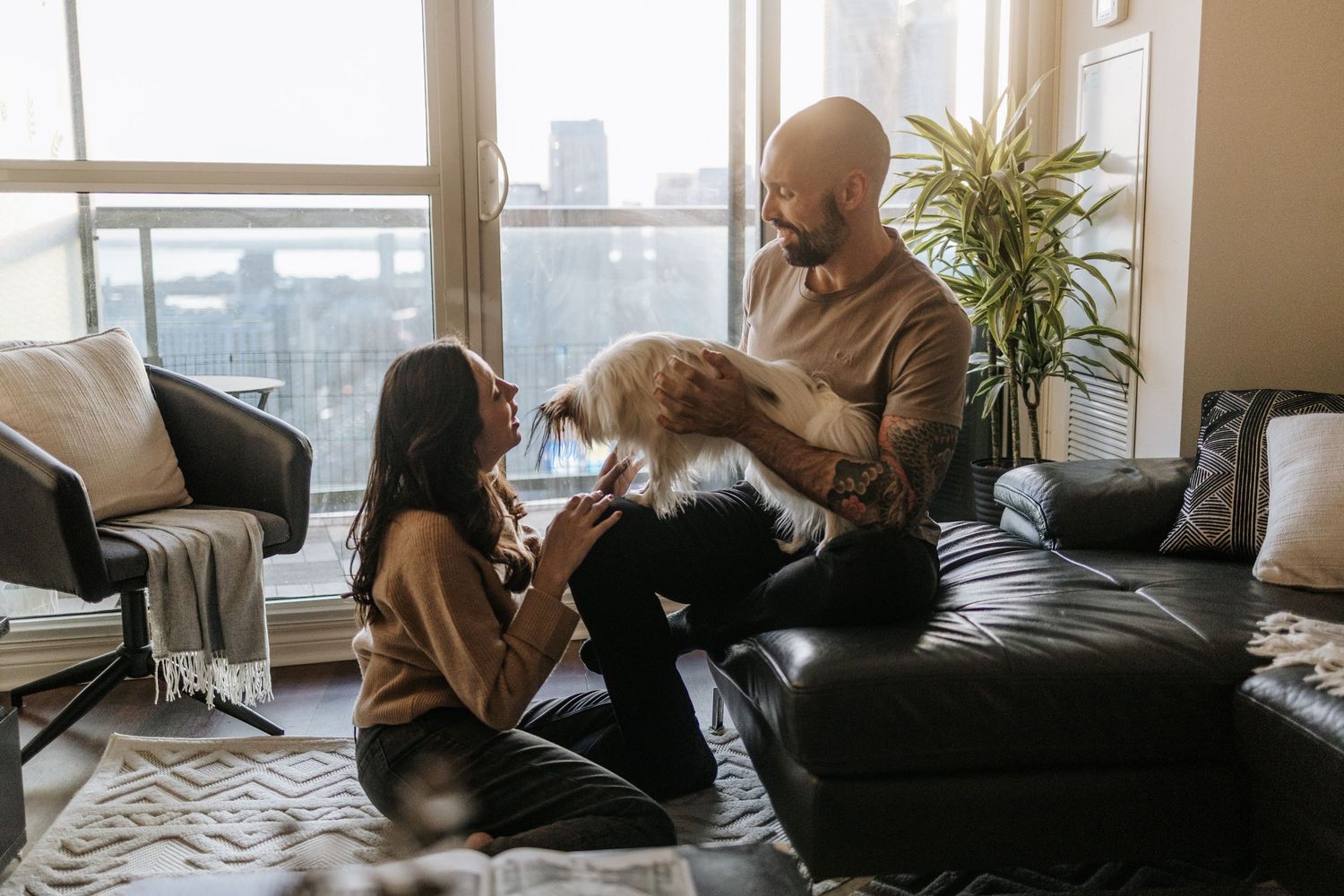 couple on the living room, the wife on the carpet, and the husband on the couch holding their dog. close to her face.