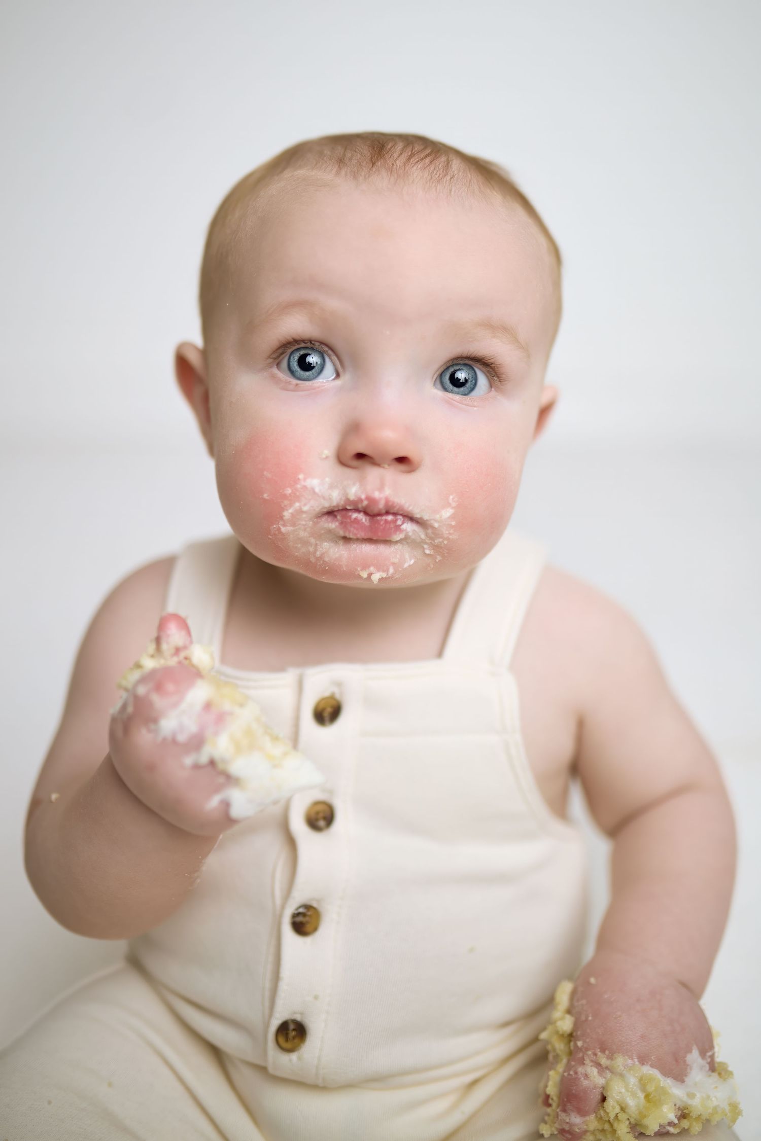 Adorable baby in cream overalls has messy face and hands from eating cake during a special photoshoot.
