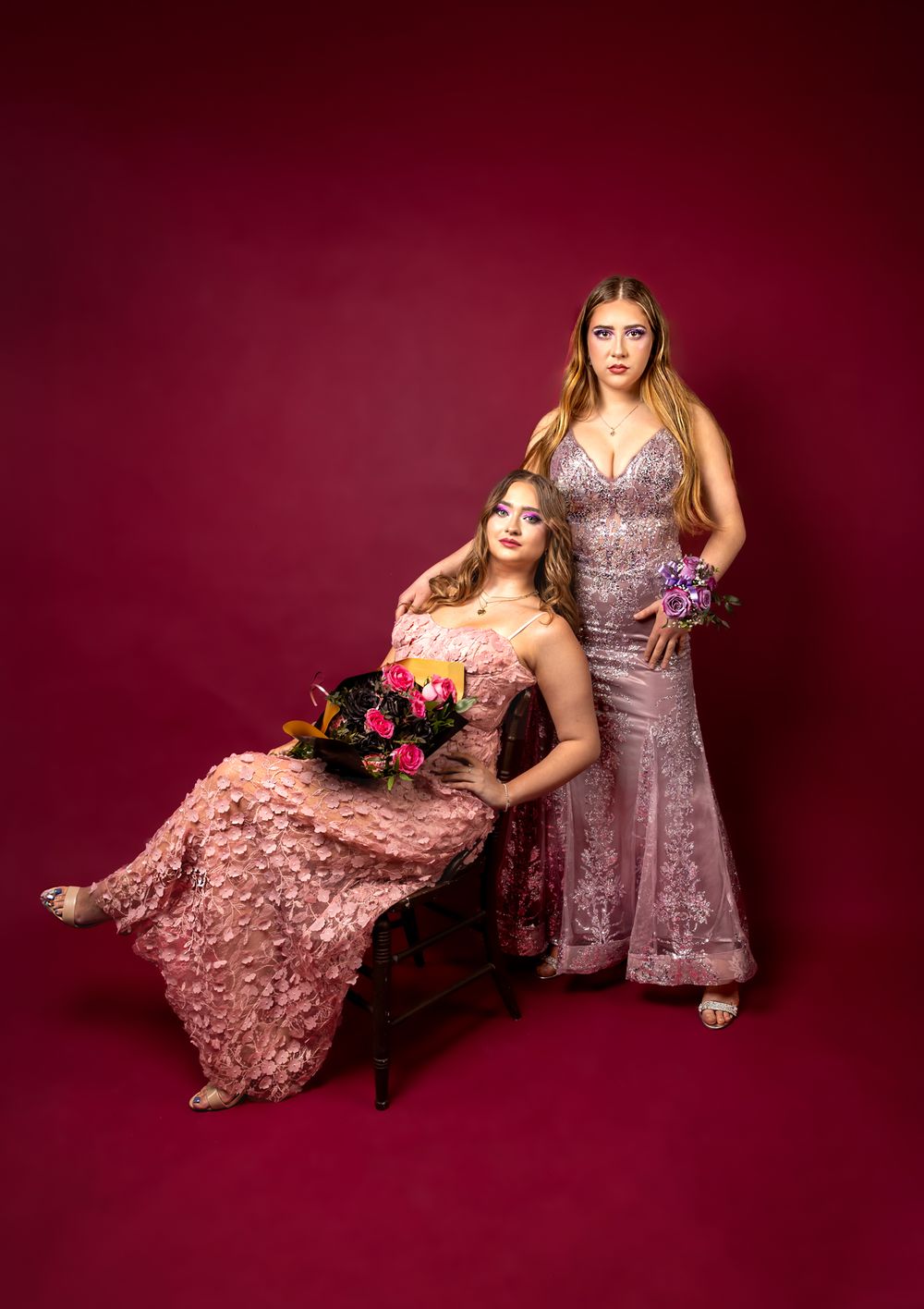Women with a pink and purple dress, matching flowers with matching makeup, in front of a red backdrop.