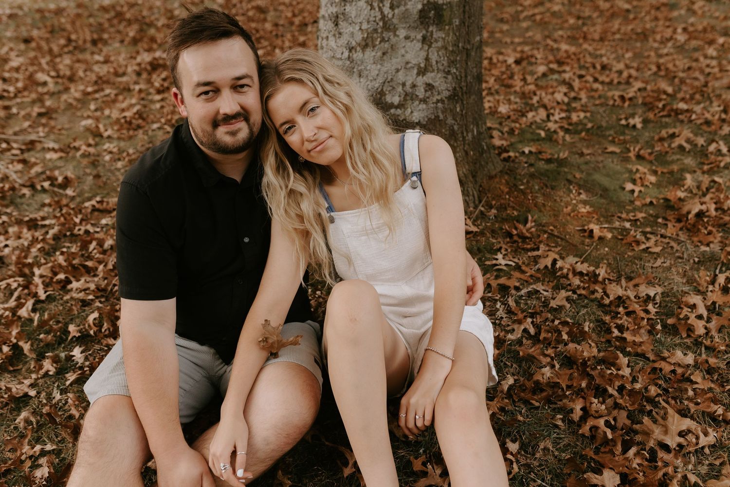 A couple sits together on fallen autumn leaves in a romantic outdoor setting.