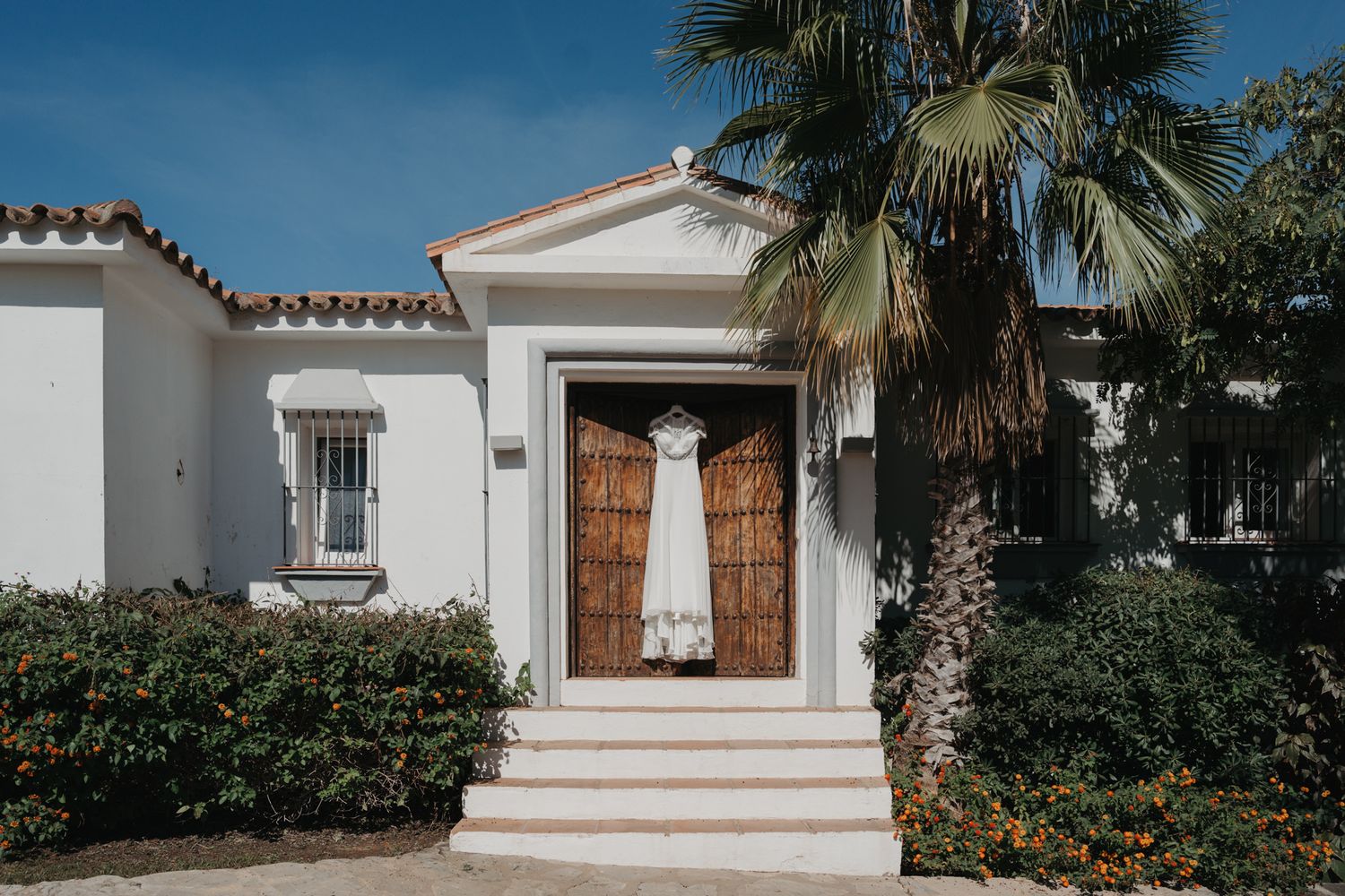 White wedding dress hanging on wooden doors of a Mediterranean-style villa with palm trees.