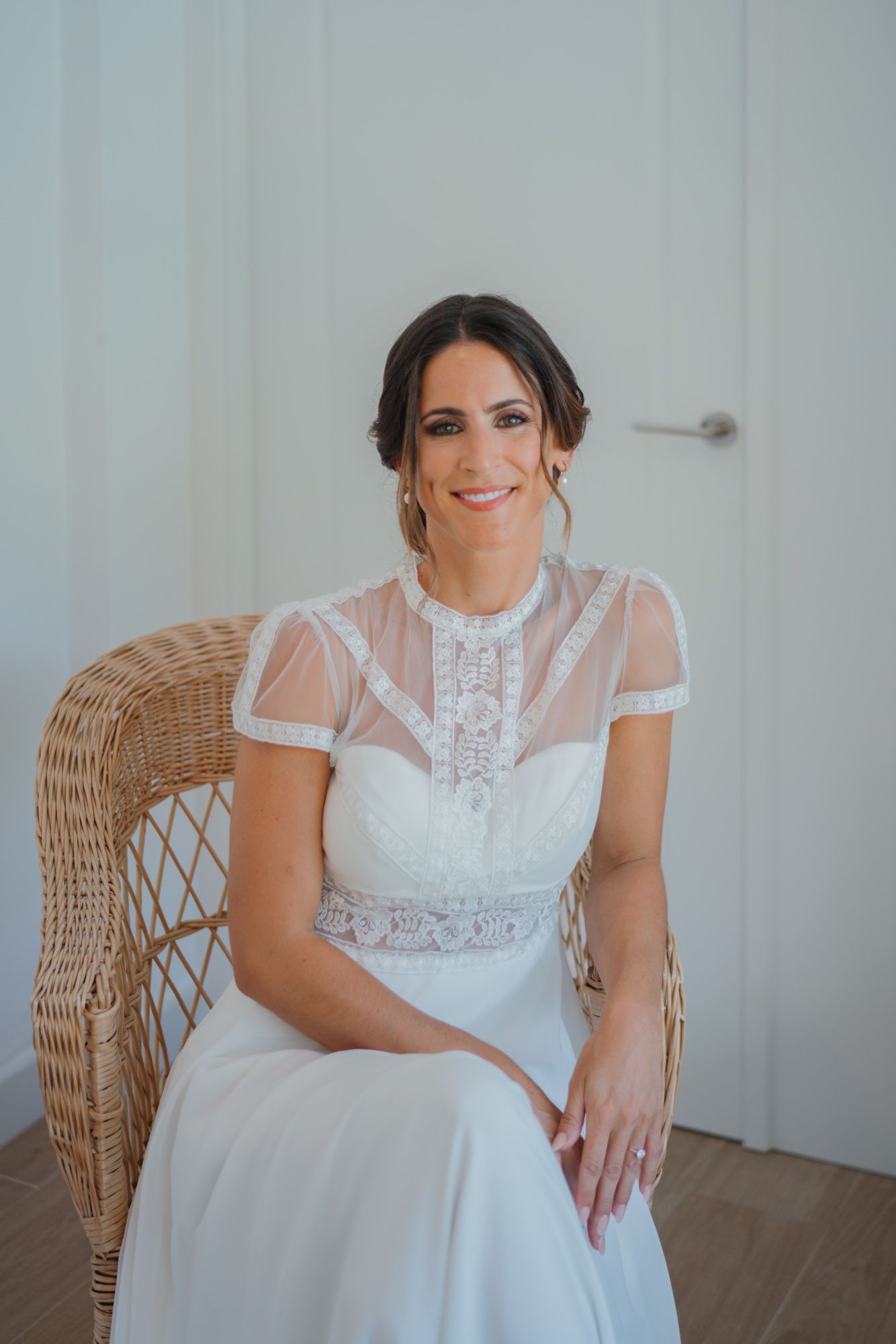 Bride in elegant white lace dress with sheer details sits in wicker chair.