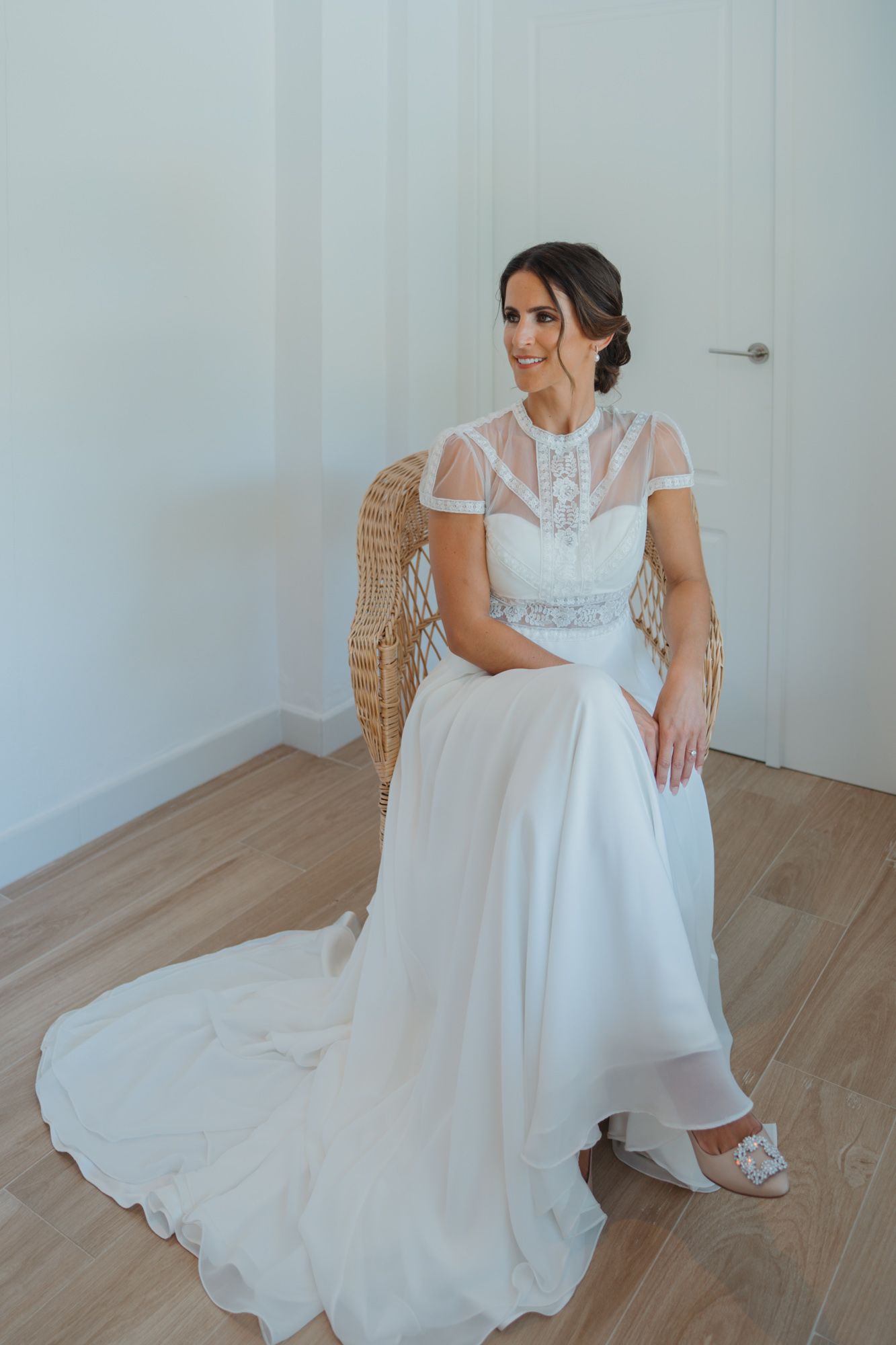 A bride in an elegant white lace gown with sheer sleeves sits gracefully in a woven chair on hardwood floors.