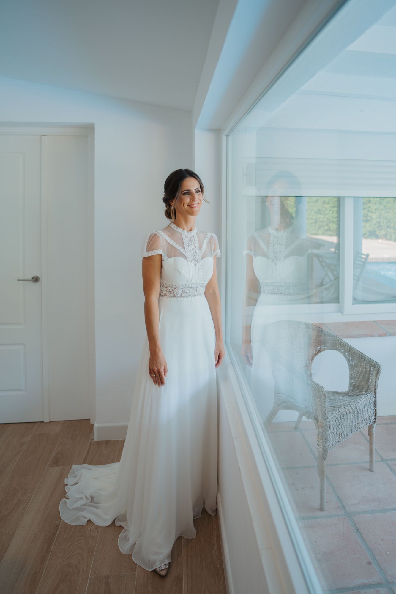 Elegant bride in lace-detailed white wedding gown stands by large window in modern interior setting.