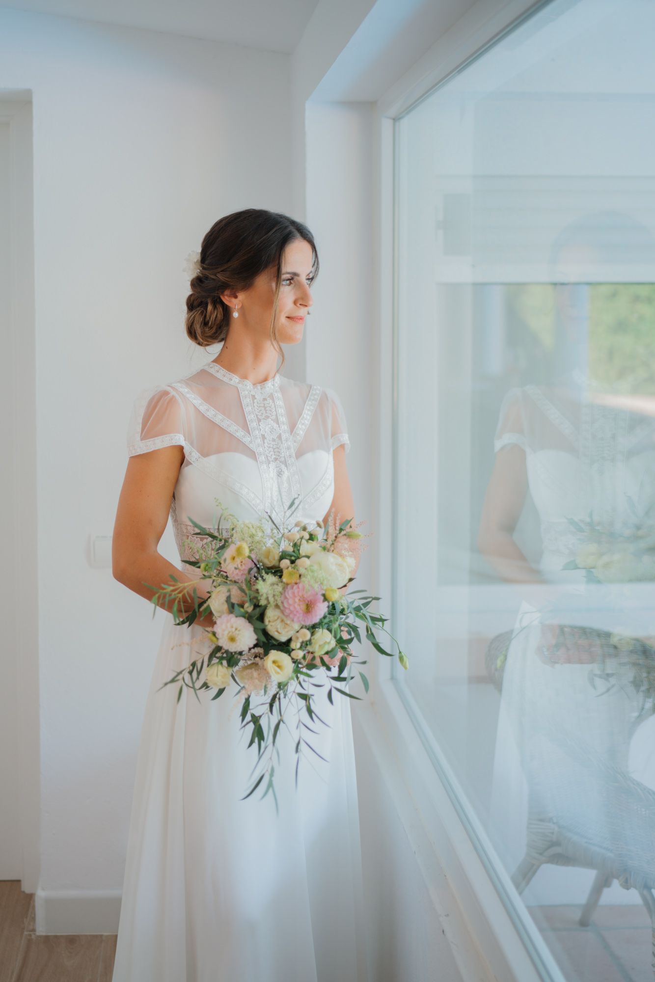 Elegant bride in lace detailed gown holding a soft pink and white bouquet by a window.