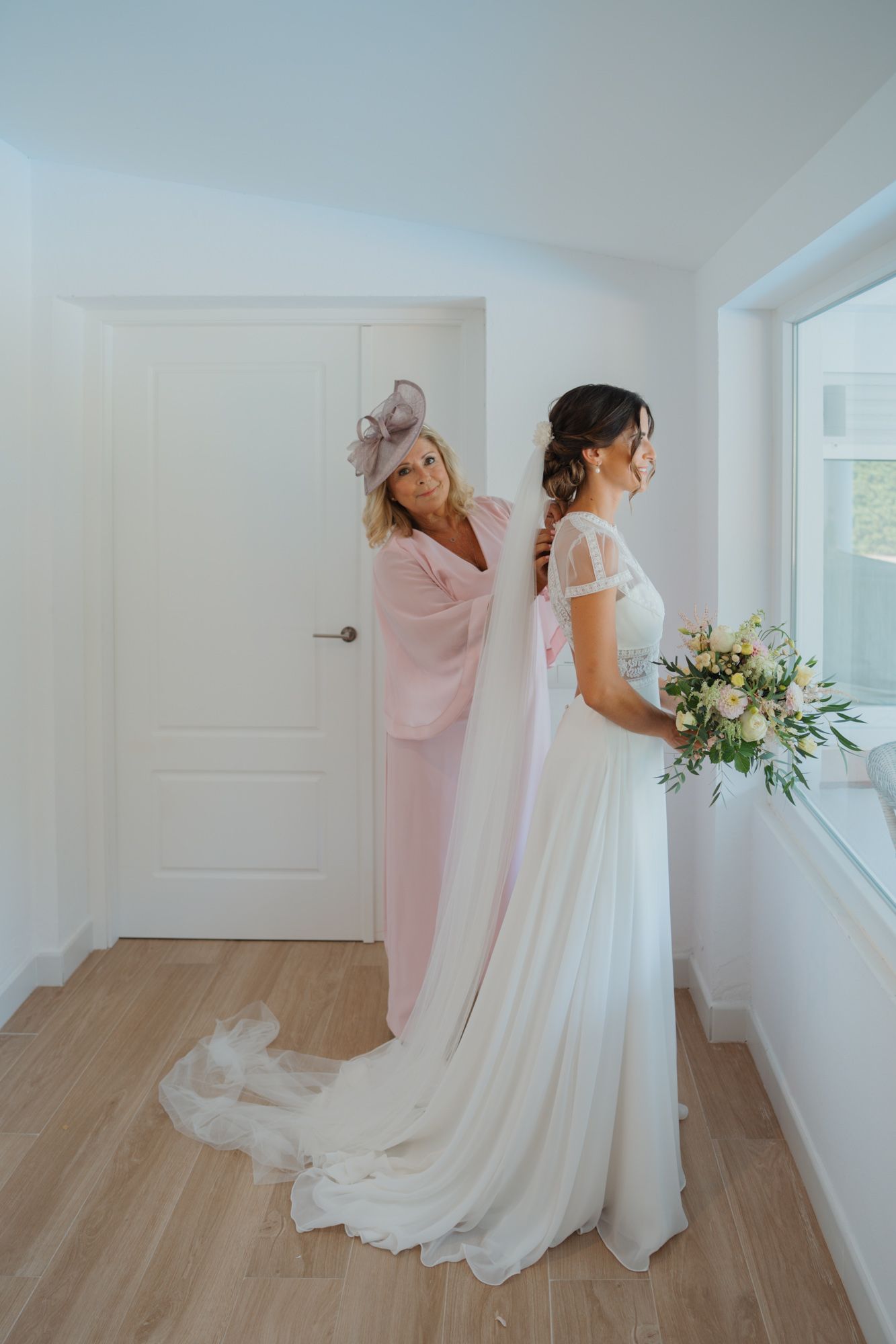 A touching moment as final wedding dress preparations are made in a bright white room.