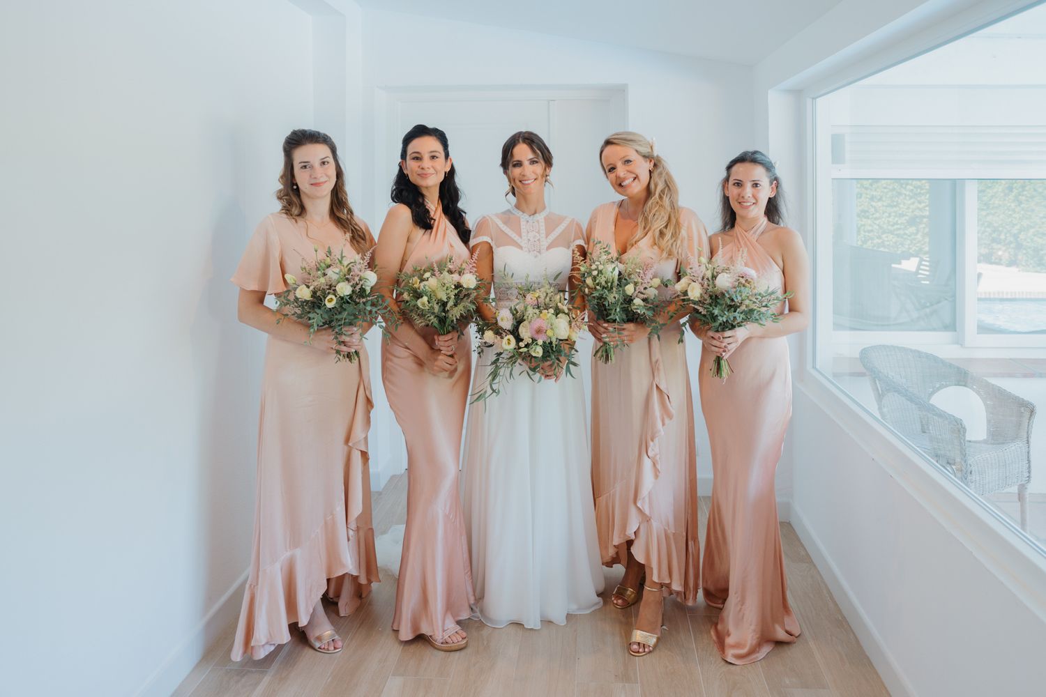 Bridesmaids in blush pink satin dresses hold elegant white floral bouquets while standing in a bright hallway.
