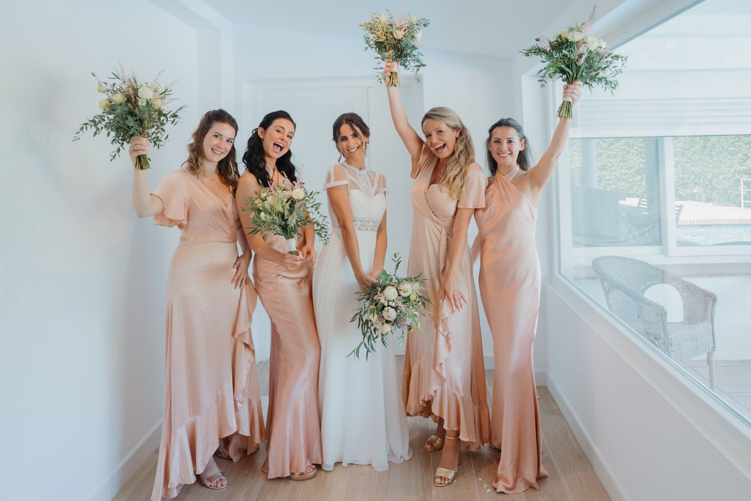 Bridesmaids in blush pink gowns pose with bouquets against white wall.