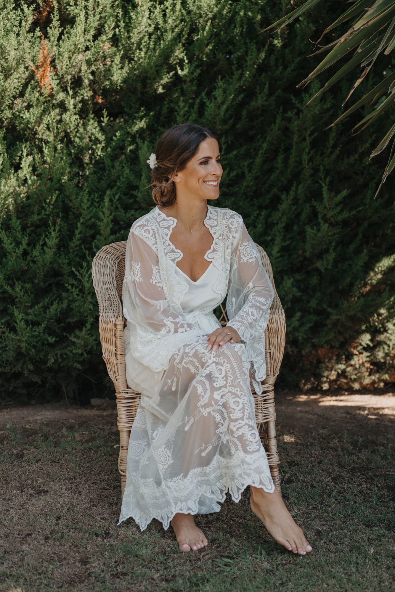 Bride in lace wedding dress sitting in wicker chair in garden setting.