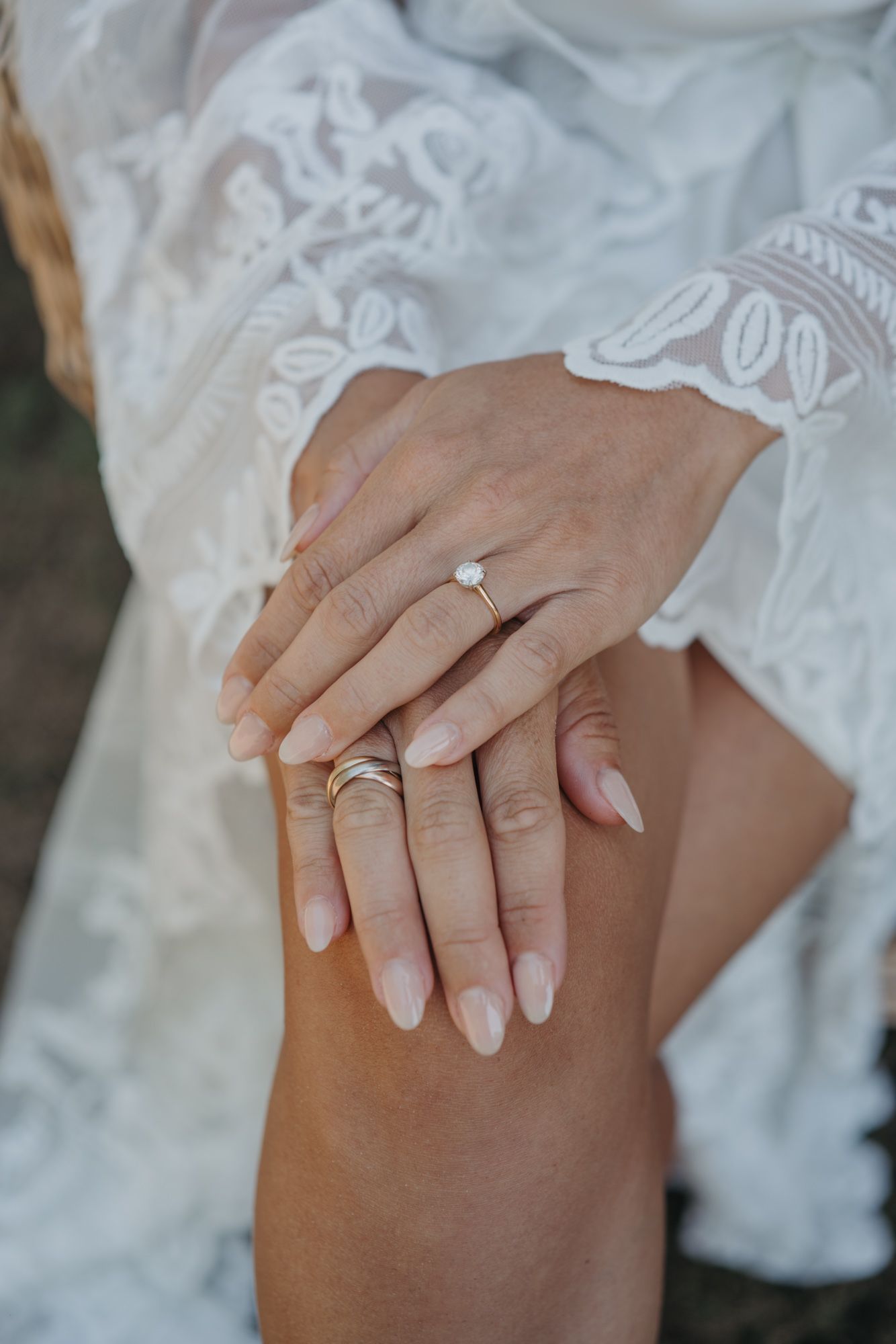 Close-up of wedding rings and manicured hands resting on white lace dress fabric.