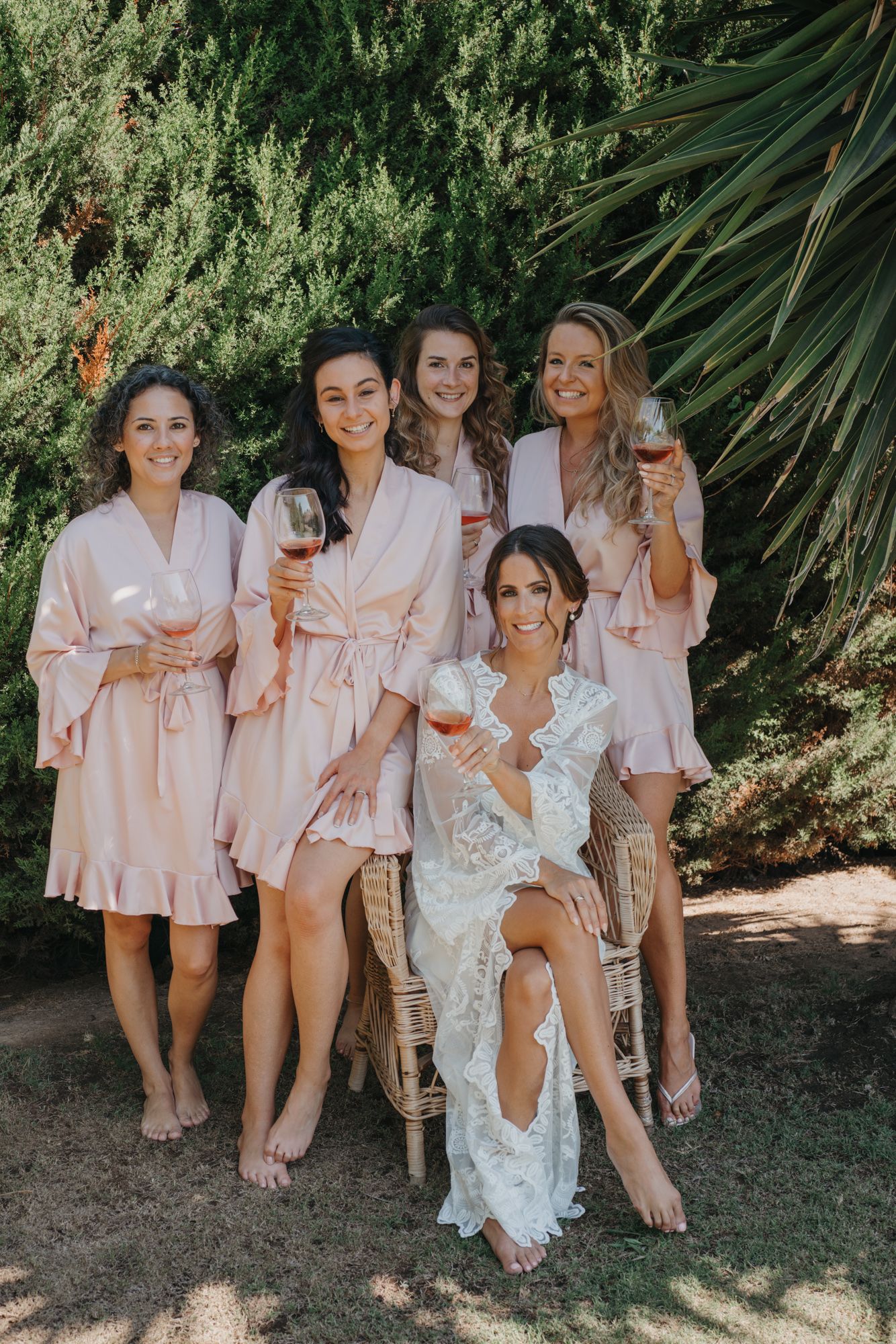 Bridal party in pink robes relaxing with wine glasses in a garden setting.