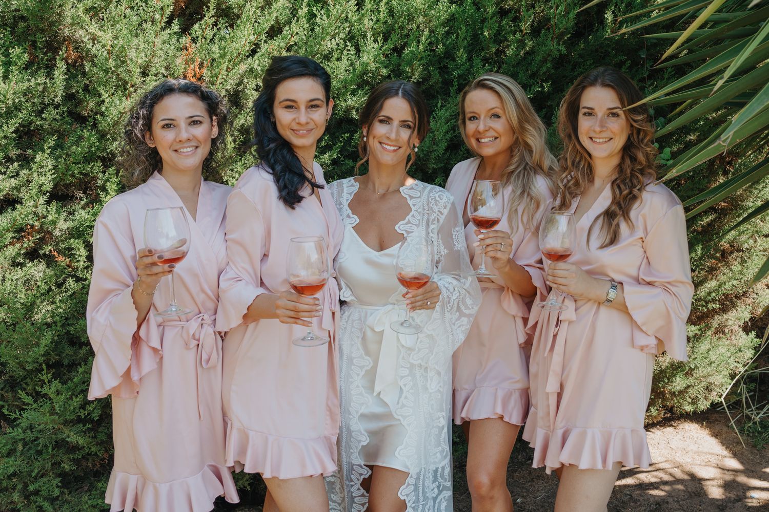 Wedding party in matching pink robes holding wine glasses against greenery backdrop.