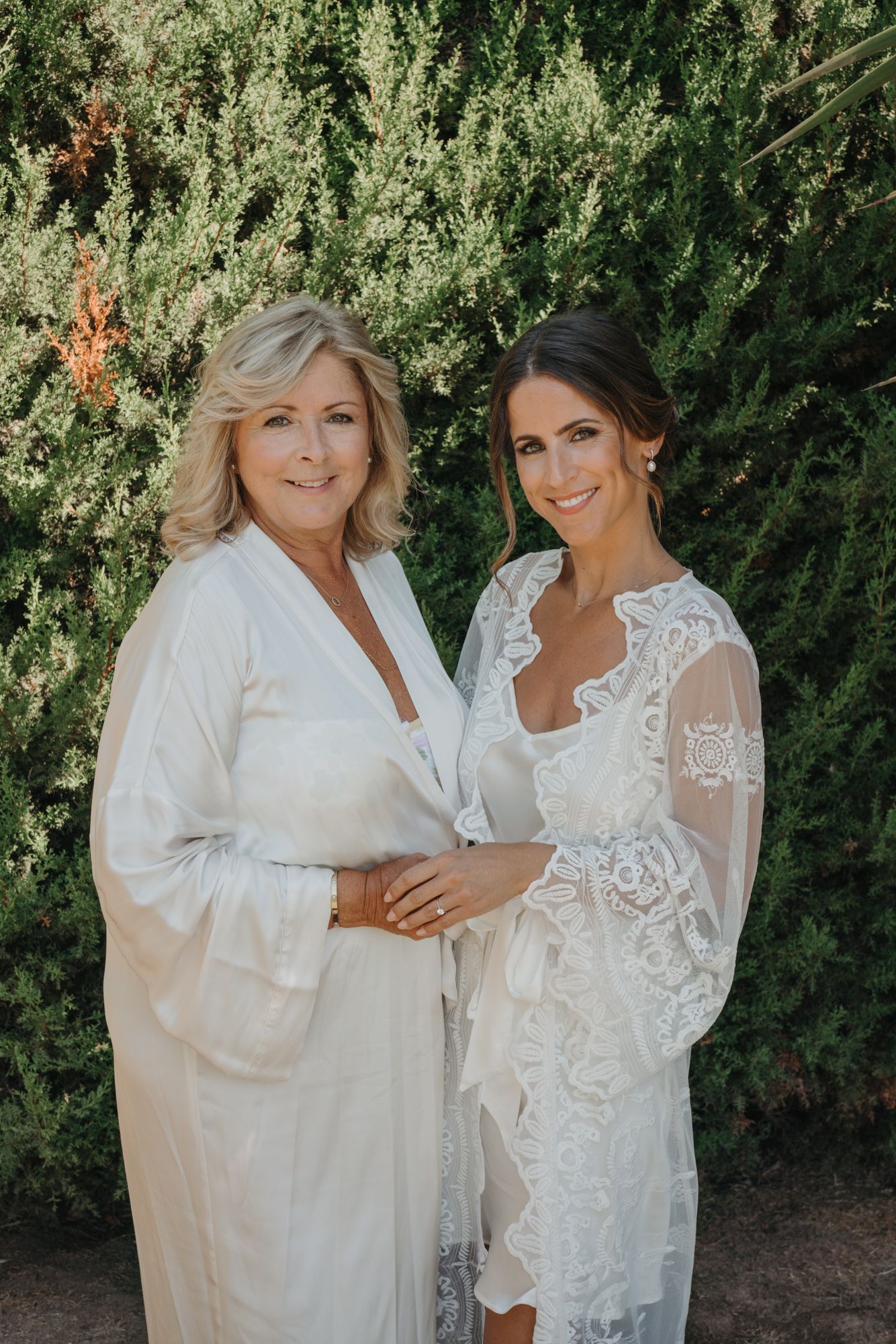 Two women in white robes pose together against a hedge backdrop.