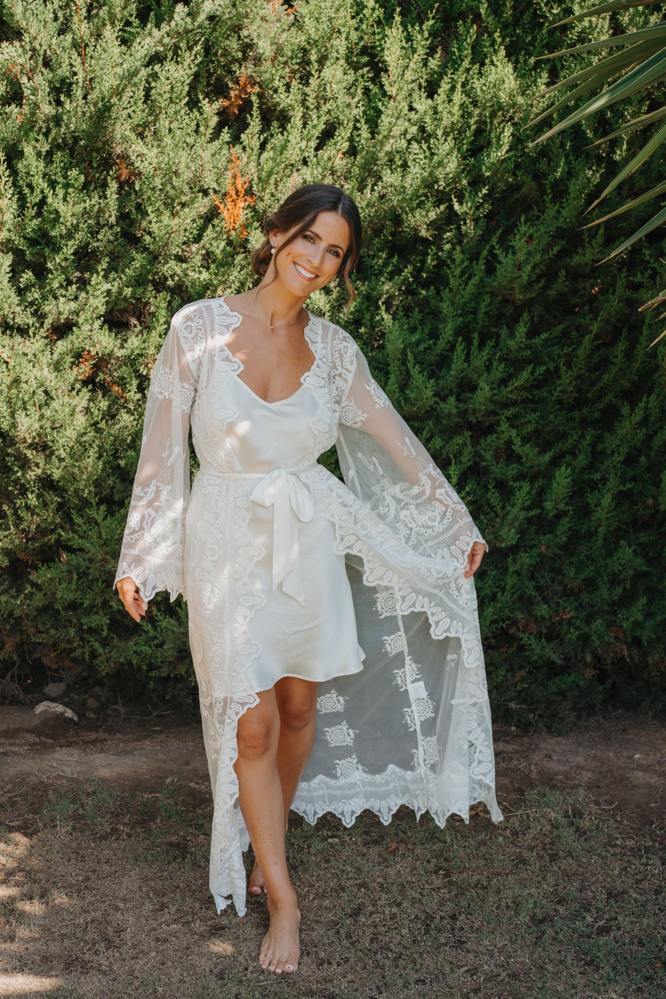 Woman in white lace robe posing barefoot against garden greenery.
