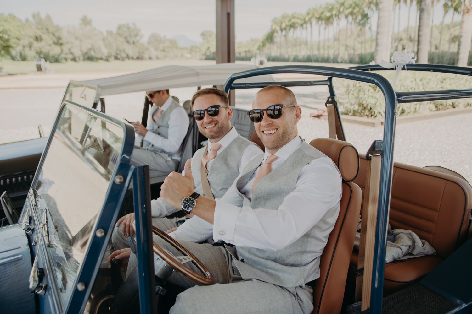 Wedding party members ride in vintage convertible car wearing stylish gray suits.