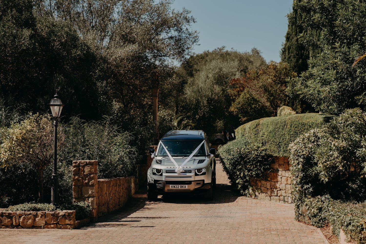 Silver wedding car driving through stone entrance with garden landscaping.