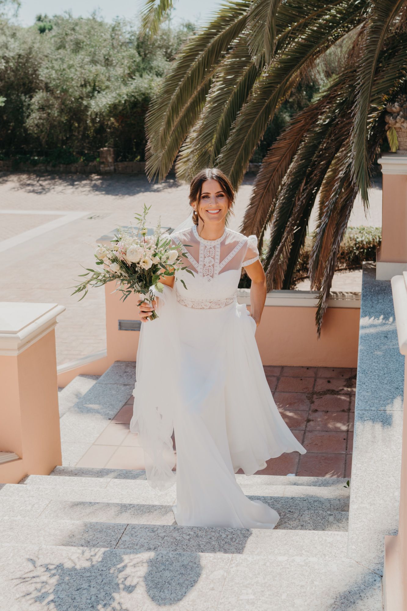 Bride holding floral bouquet poses on sunny steps under palm trees.