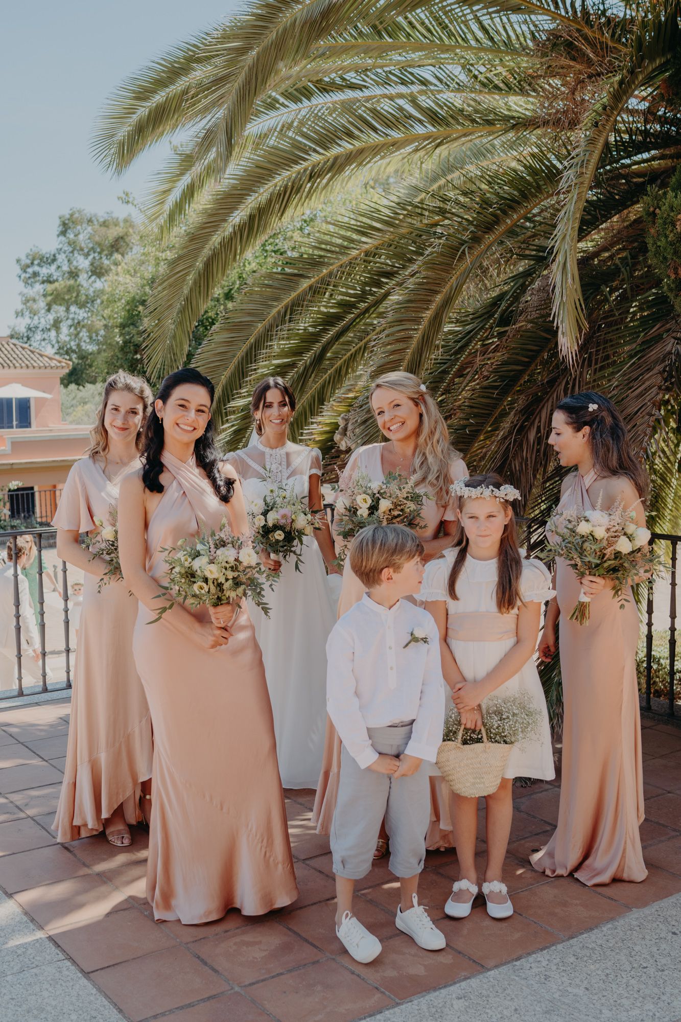 Wedding party in blush pink dresses poses under a palm tree.