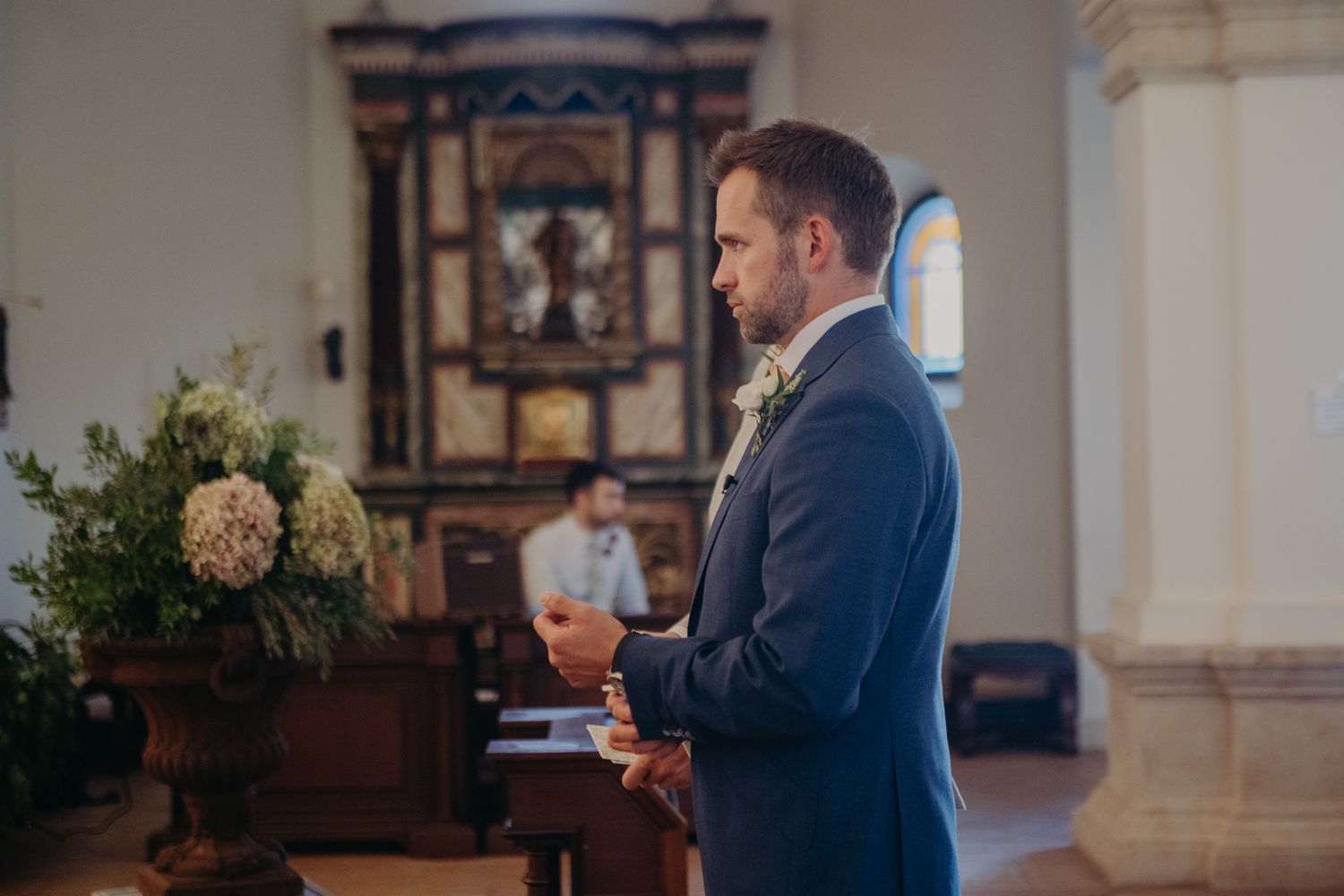 Groom in navy suit holds drink while standing in ornate church interior.