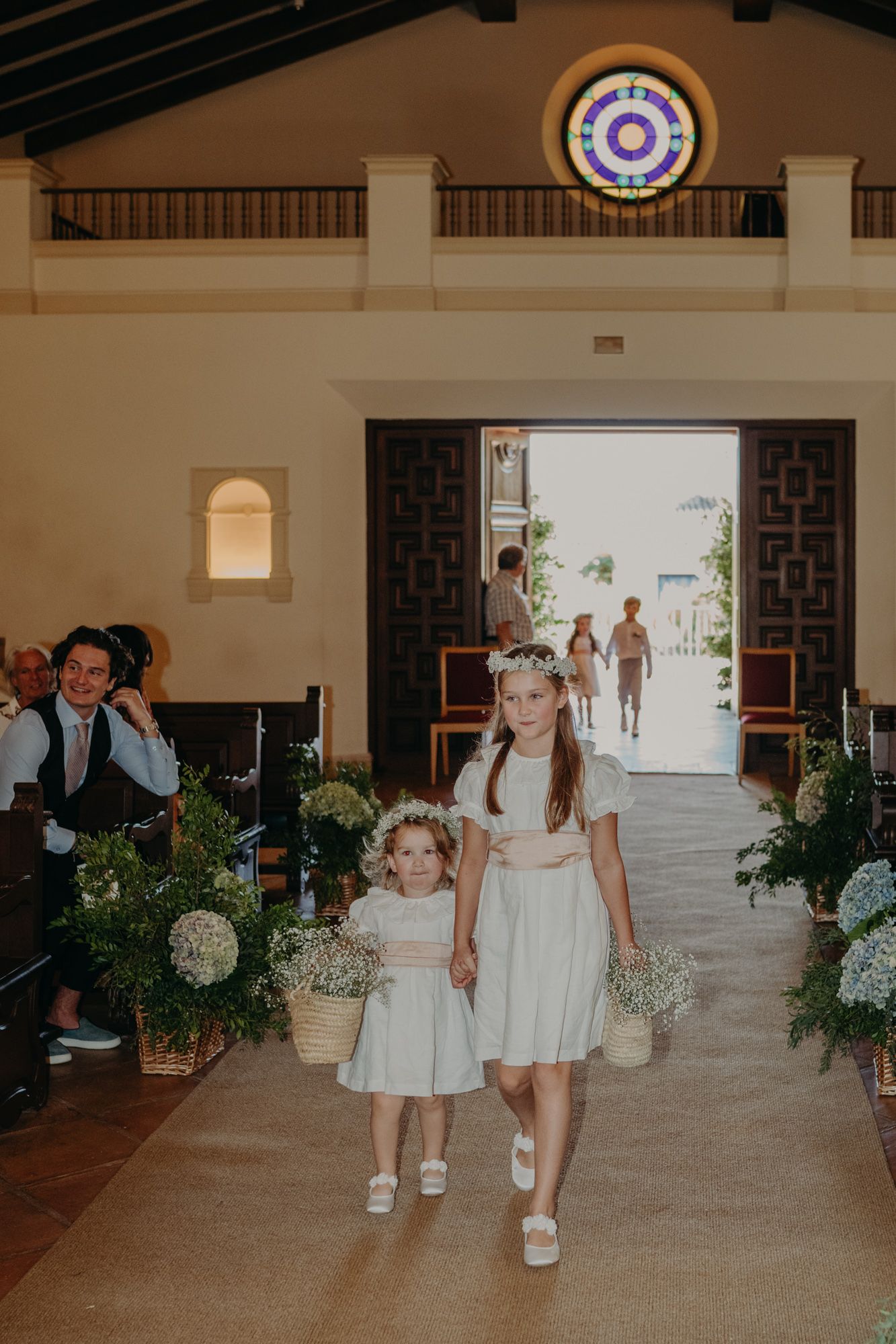 Two young attendants in white dresses walk down a church aisle adorned with floral arrangements.