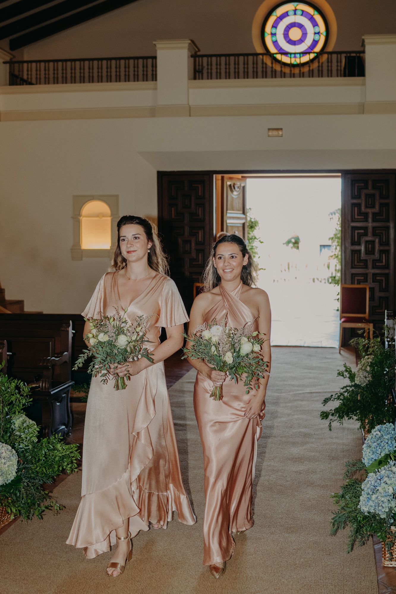 Two attendants in blush silk dresses walk down an indoor aisle carrying greenery bouquets.
