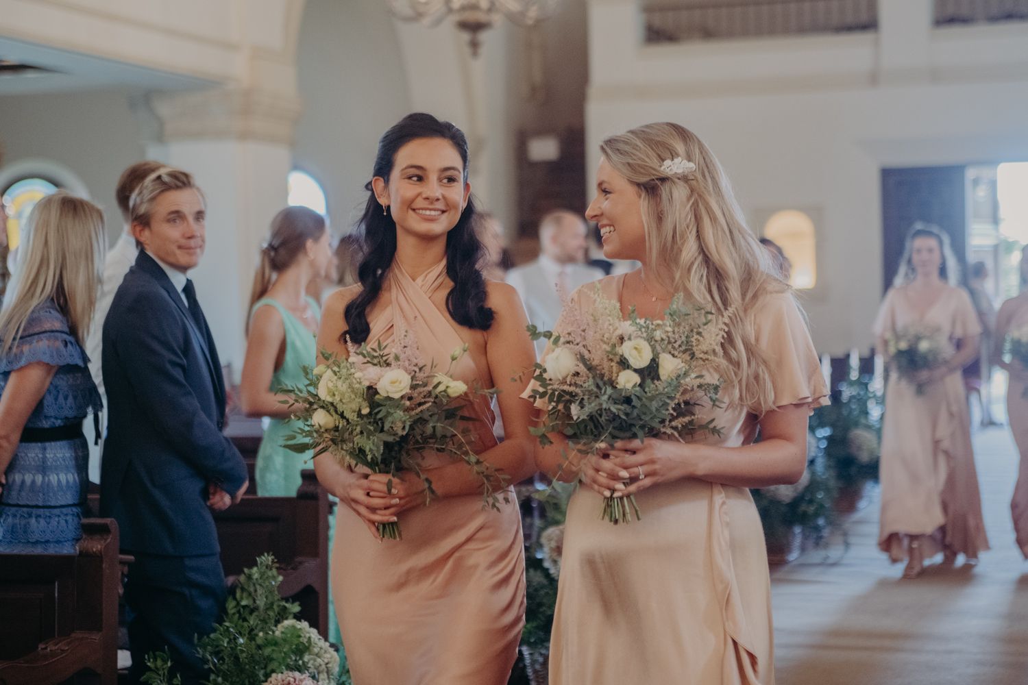 Bridesmaids in blush satin dresses carrying bouquets walk through elegant church interior.