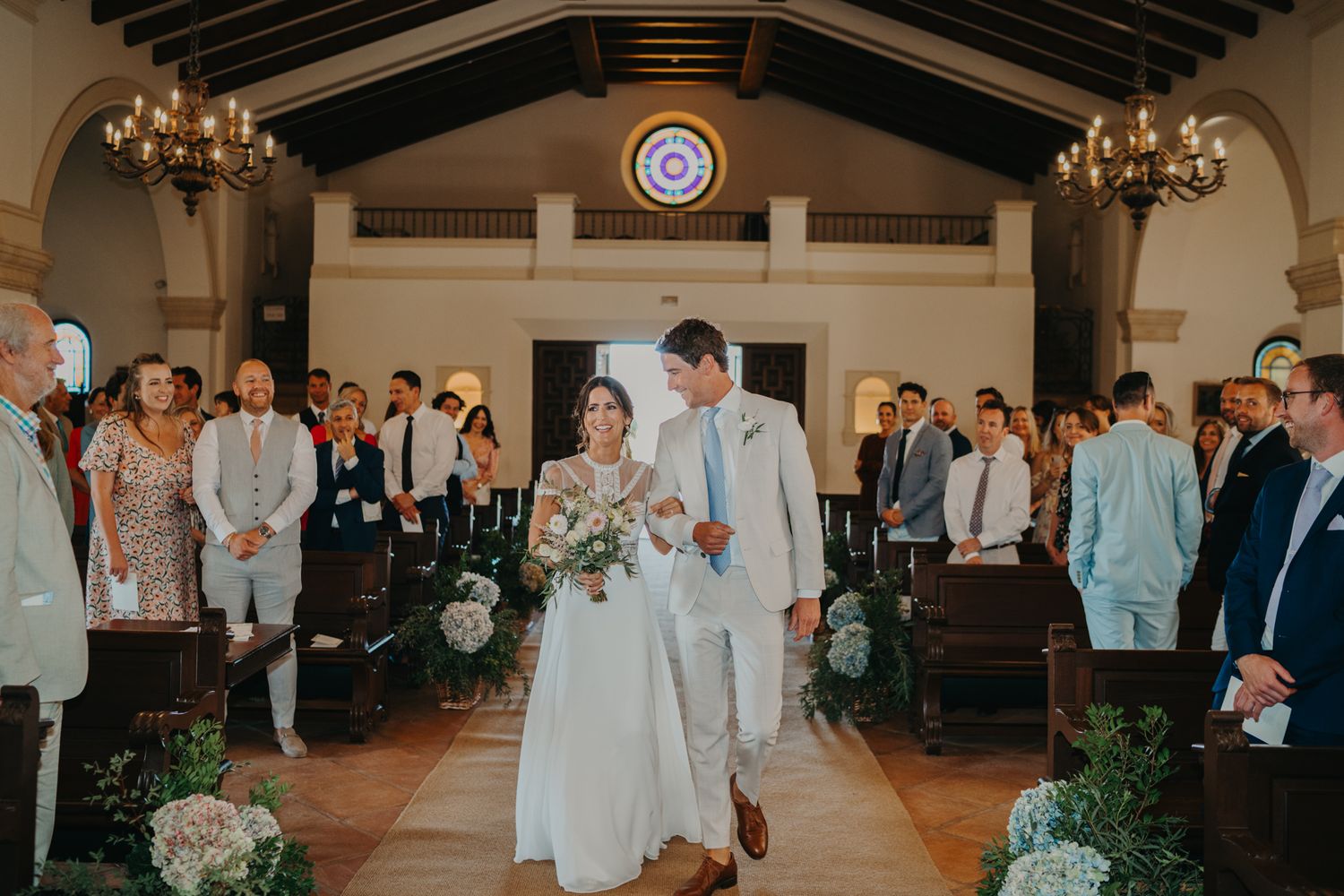 A lovely wedding ceremony takes place in a chapel with wooden beams and chandeliers as guests look on.