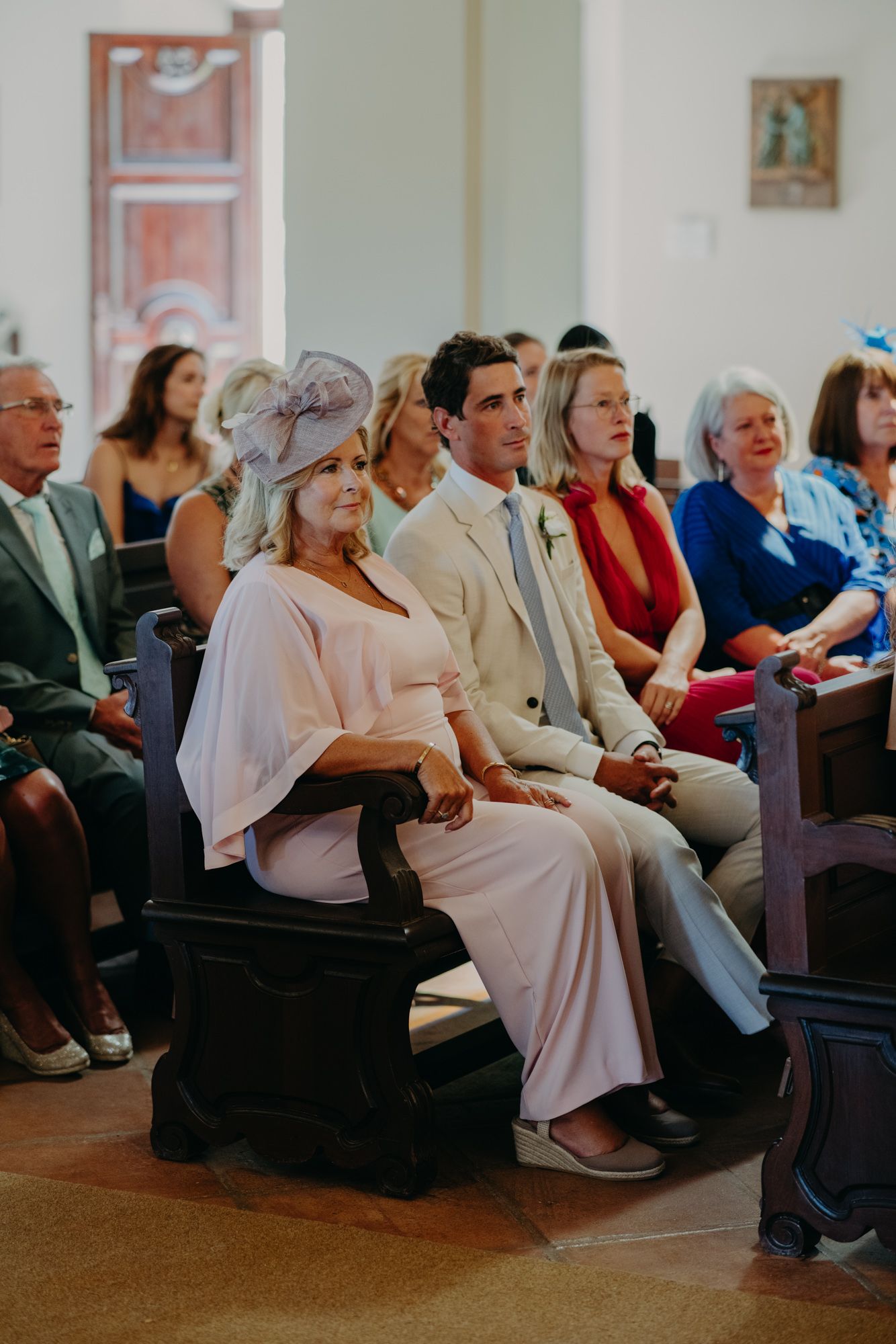 Wedding guests seated in church pews wearing formal attire and summer dresses during a ceremony.