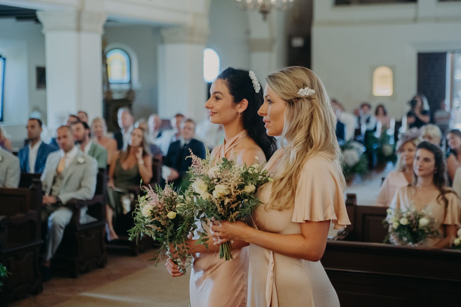 Bridesmaids stand together in a church holding elegant green and white floral bouquets during a wedding ceremony.