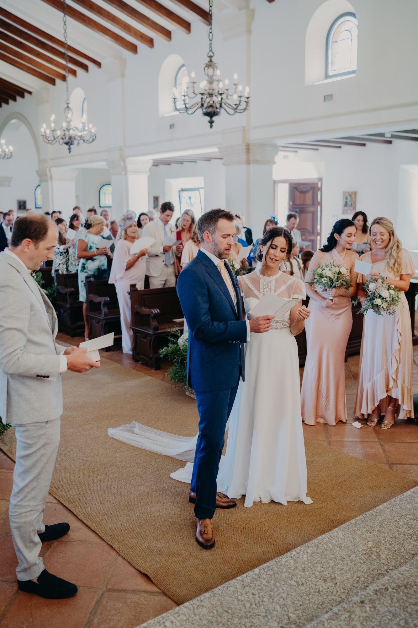 Wedding ceremony in elegant white church interior with chandeliers and gathered guests.