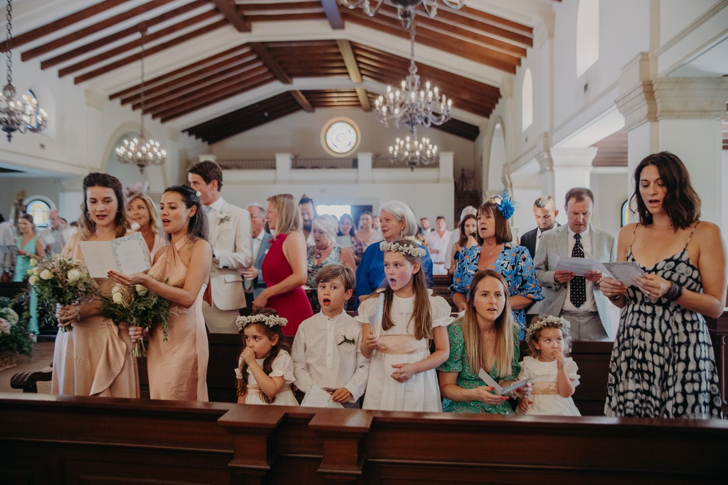Wedding guests sit in church pews during ceremony with elegant chandeliers and exposed wooden beams overhead.