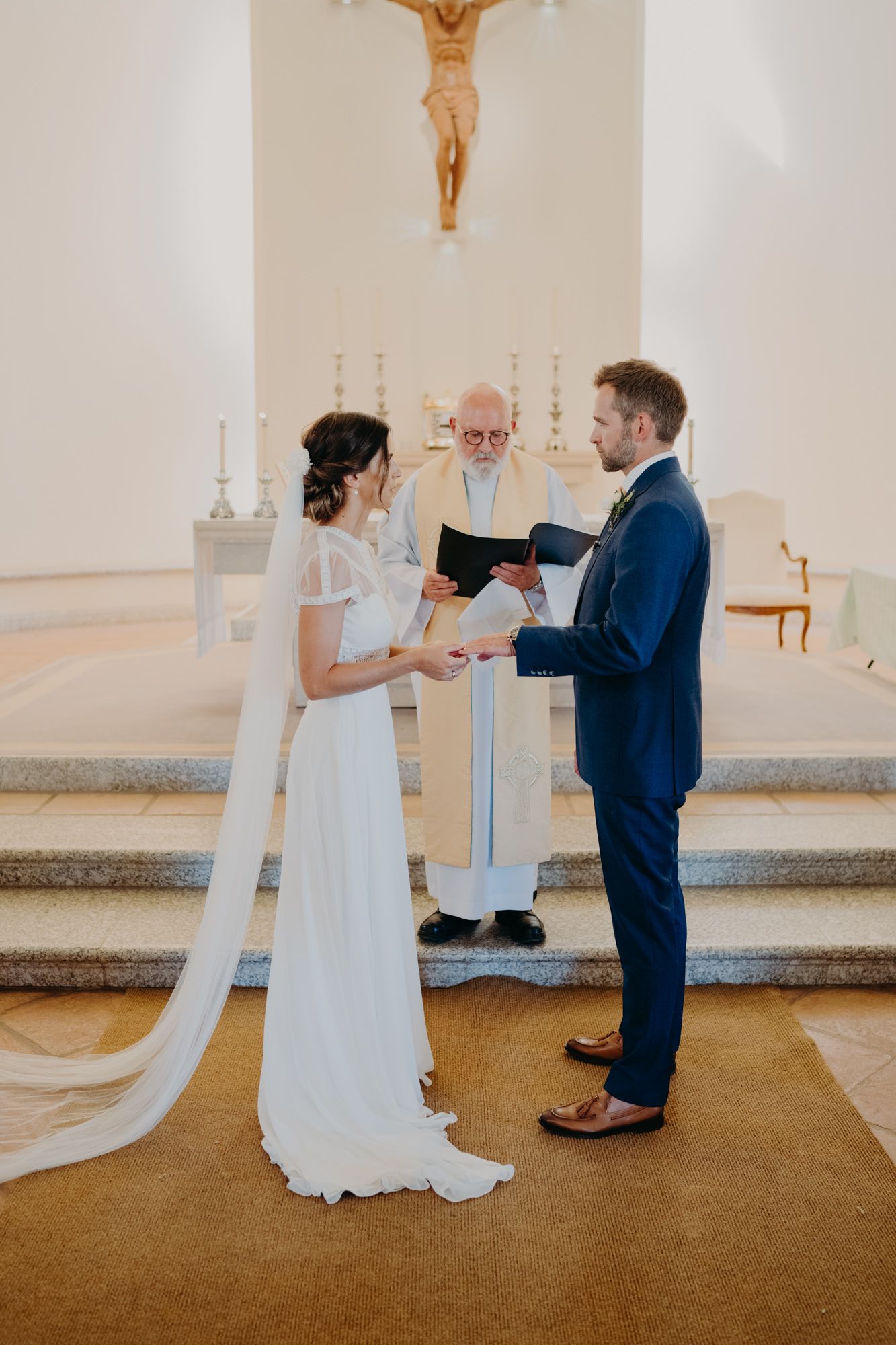 A traditional church wedding ceremony with couple exchanging vows before a priest under a prominently displayed crucifix.