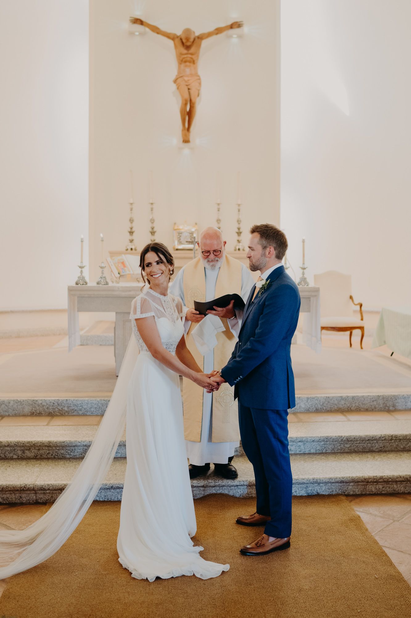 Couple holds hands during their wedding ceremony in a church with a crucifix and altar in the background.