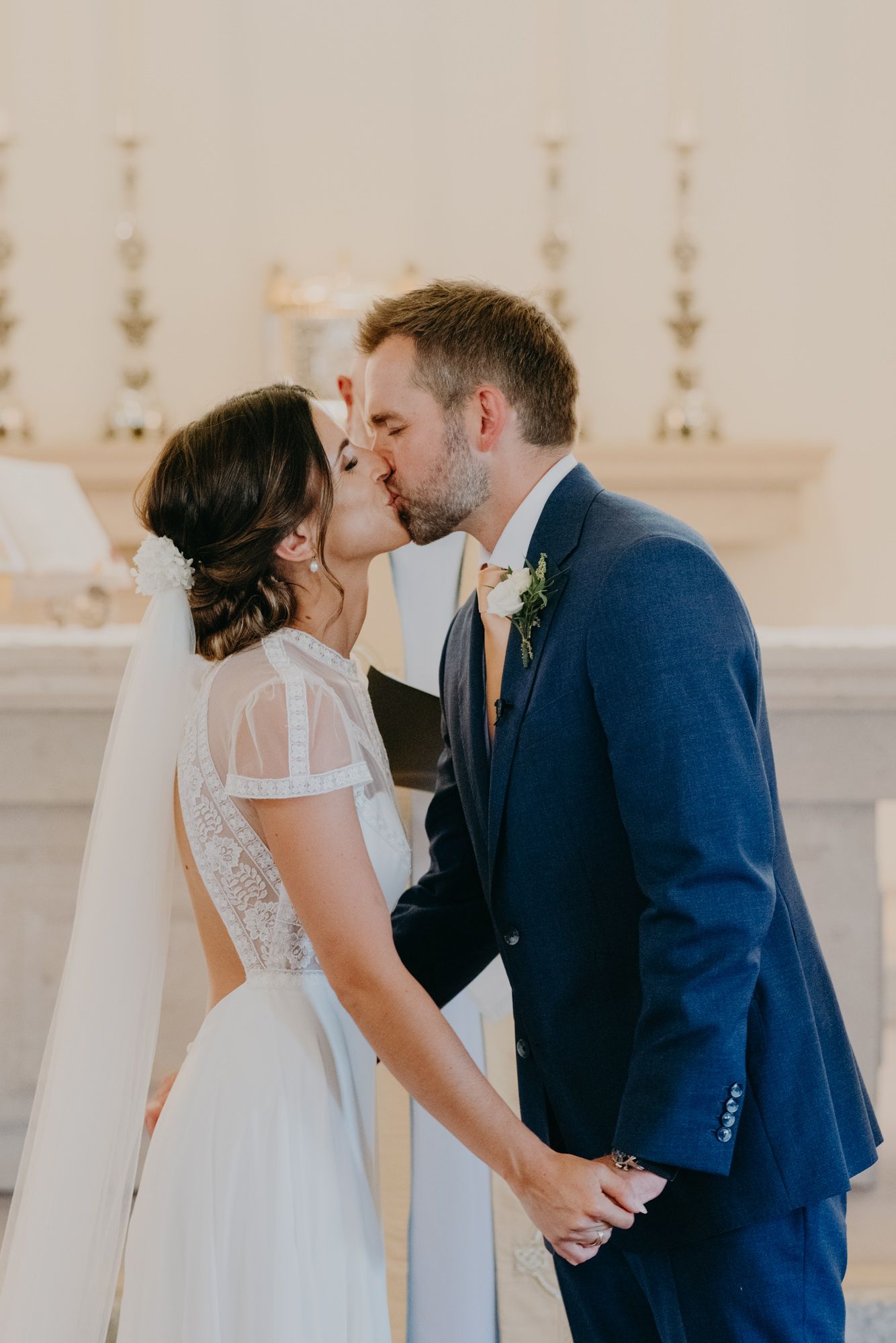 A romantic wedding kiss between bride in lace gown and groom in navy suit at elegant white altar.