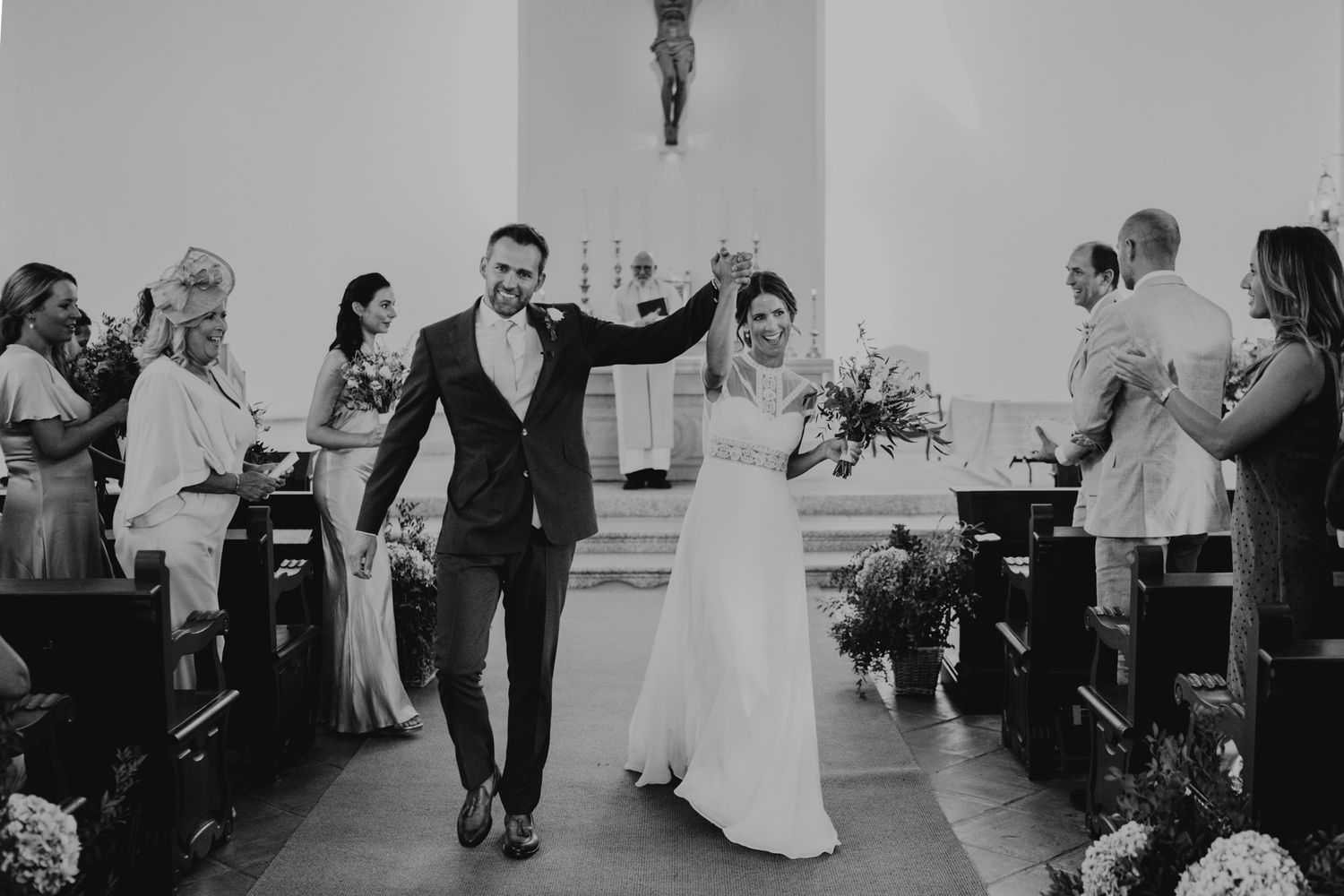 Newlyweds joyfully exit church ceremony aisle while guests celebrate in black and white photo.