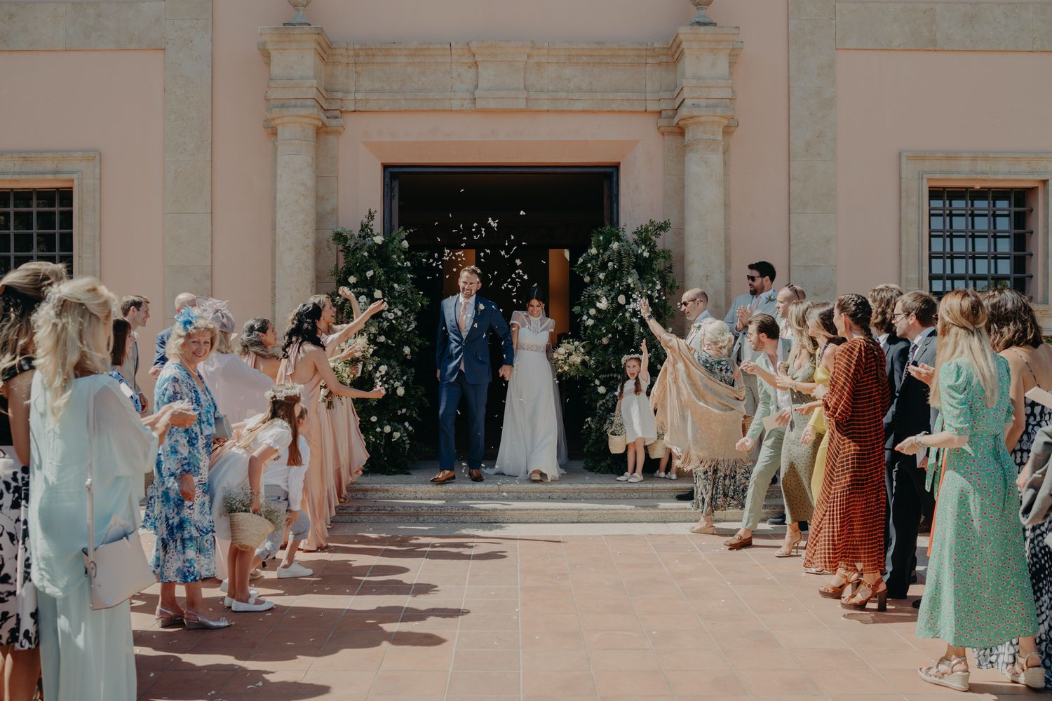 Wedding ceremony exit with guests lining a walkway outside a pink Mediterranean-style building.