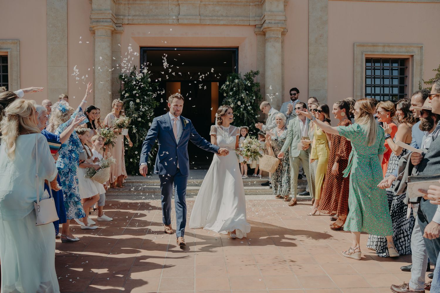 Wedding celebration outside a classic building as guests throw flower petals during a joyful exit.