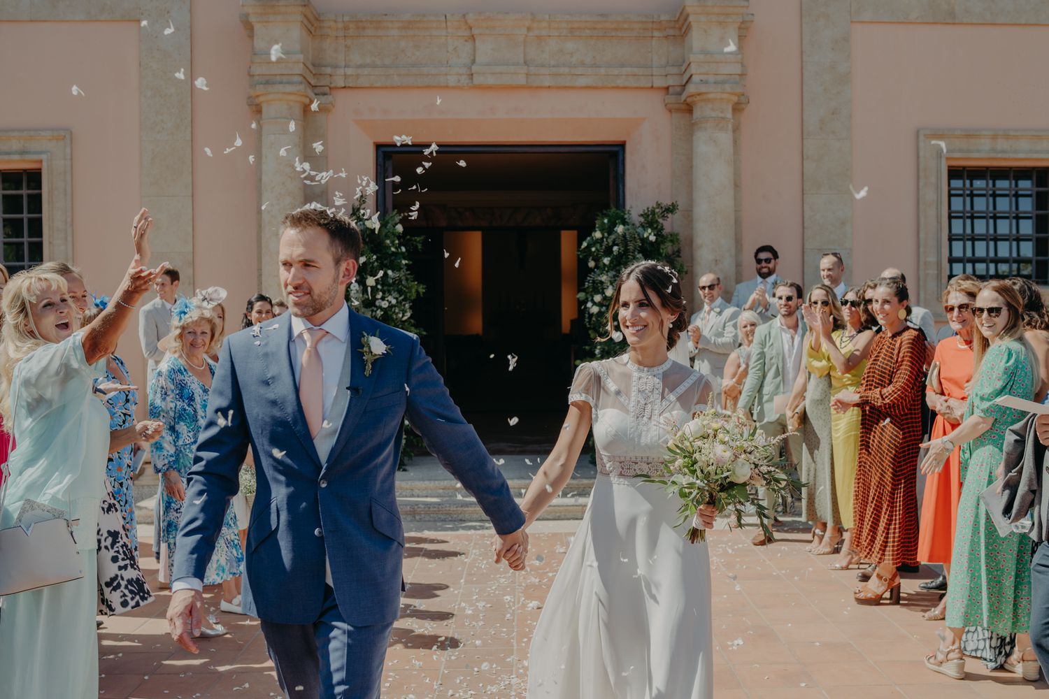 Newlyweds exit their wedding ceremony while guests throw confetti outside a pink building.