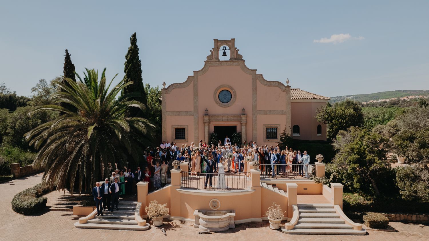 Spanish-style church exterior with palm trees and wedding guests gathered on steps.