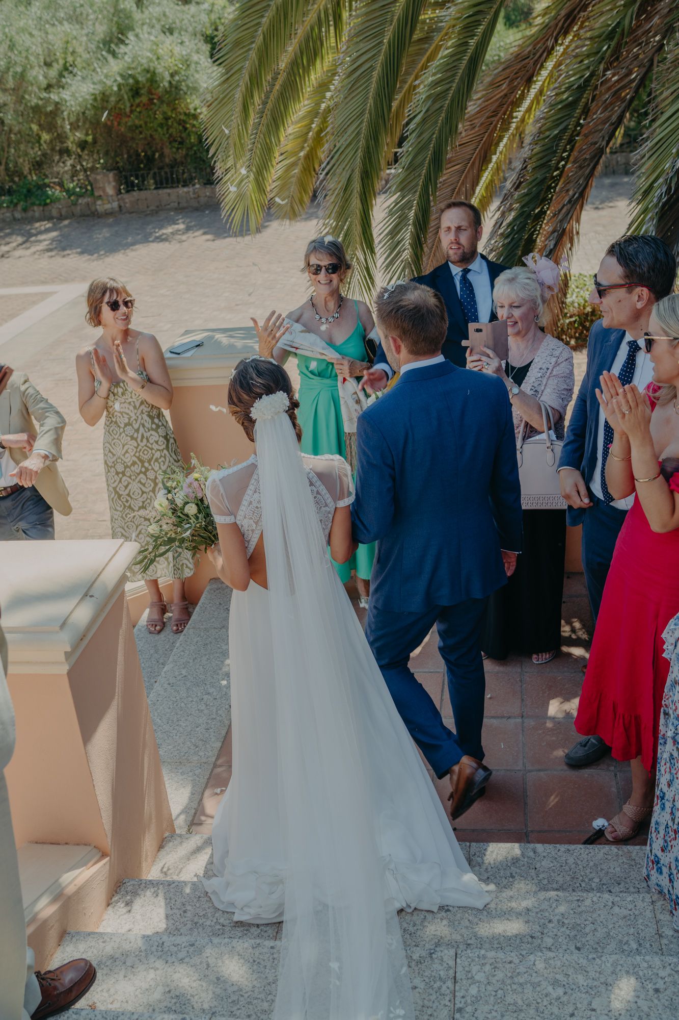 Wedding guests gather on a sunny Mediterranean terrace surrounded by palm trees.